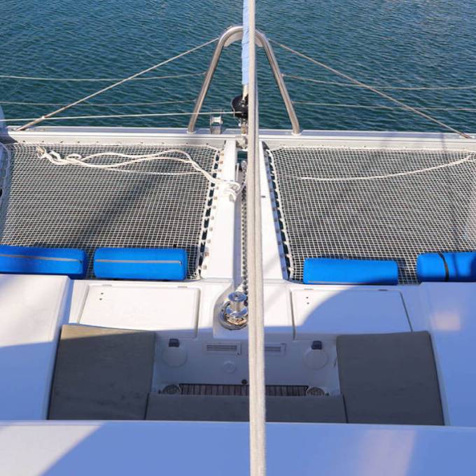 Top view of a catamaran bow with nets and blue cushions over calm water.