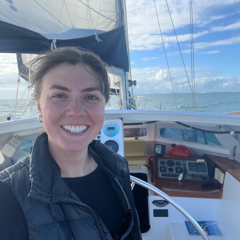 Person smiling on a sailboat with blue sky and ocean in the background.