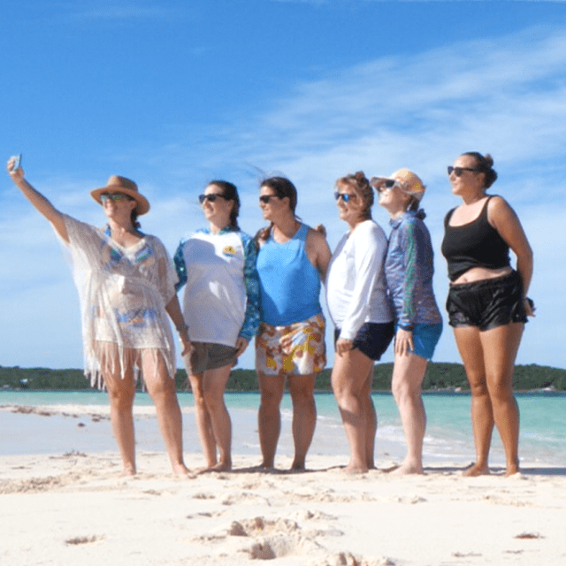 Six people posing for a selfie on a sandy beach under a clear blue sky.