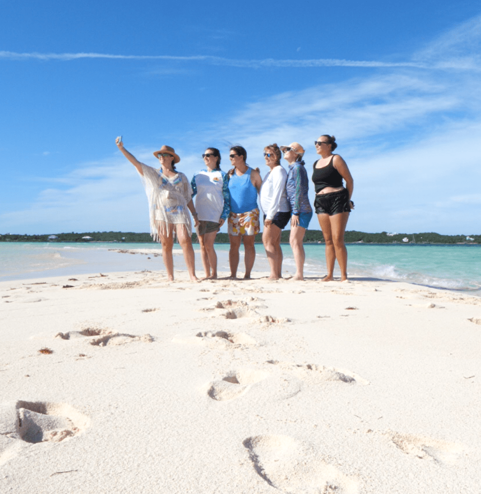 Six people posing for a selfie on a sandy beach under a clear blue sky.