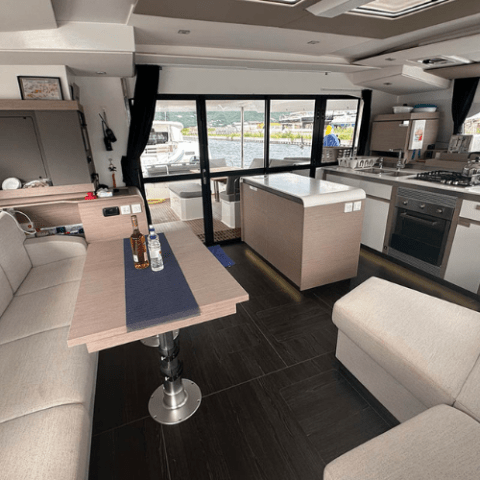 Spacious yacht interior with sofas, table, kitchen, and large windows showing marina view.