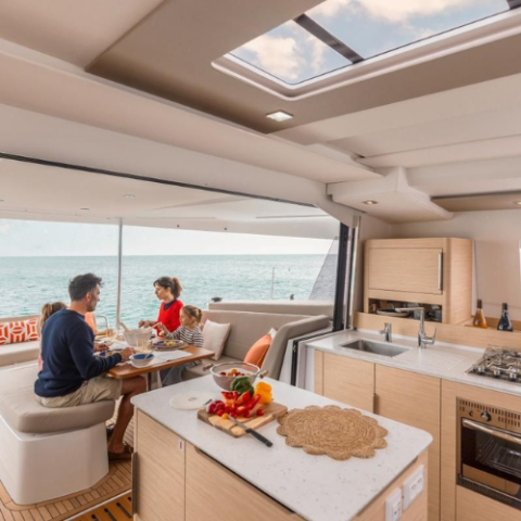 Family dining inside a yacht, with an ocean view through large windows and a modern kitchen visible.