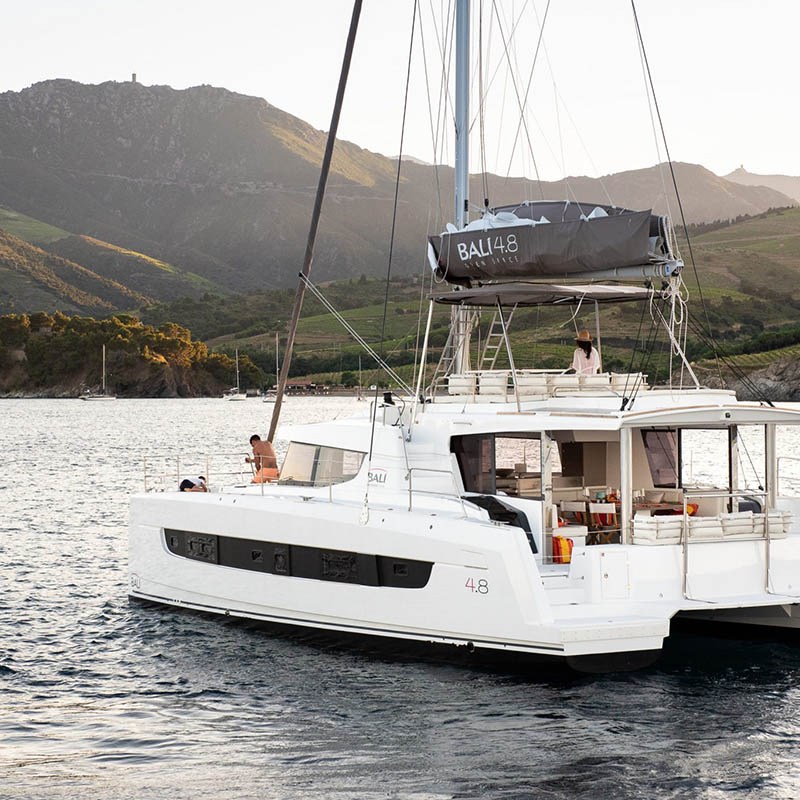 A white catamaran sails near a mountainous coastline under a clear sky.