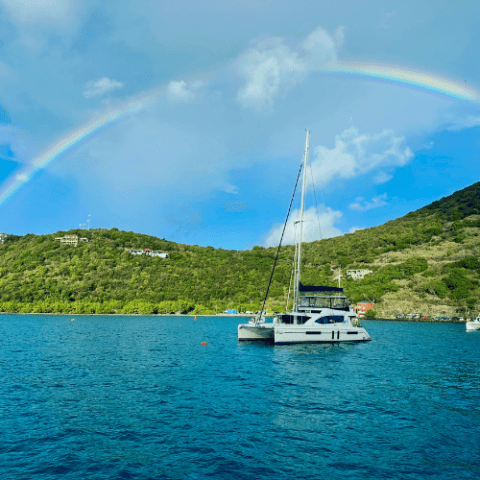 Sailboat on blue water with rainbow and green hills in background.