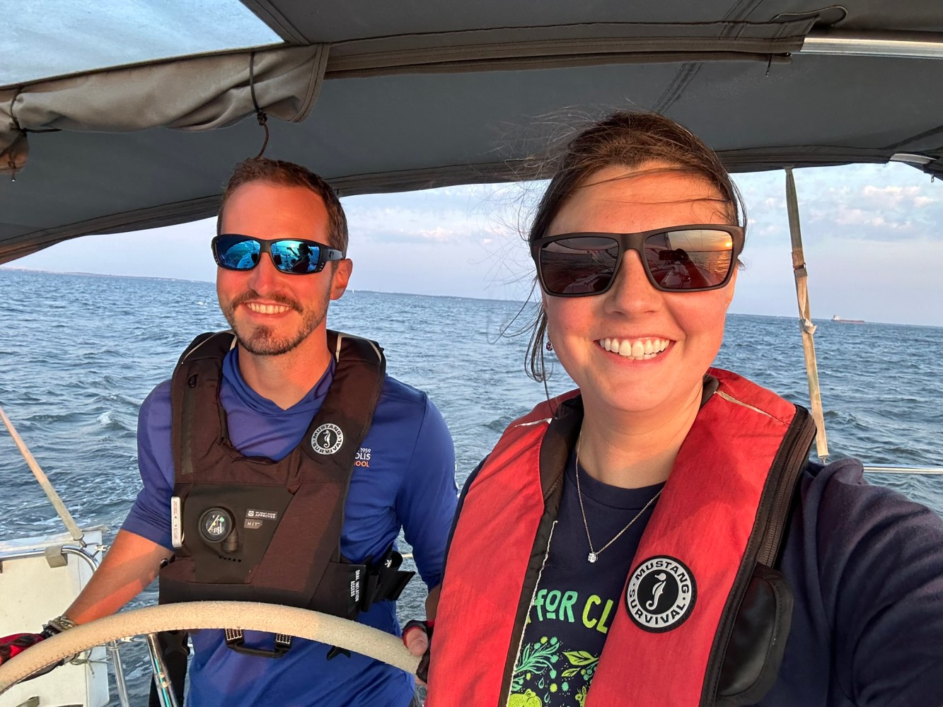 Two people wearing sunglasses and life jackets smiling on a boat, with ocean in the background.