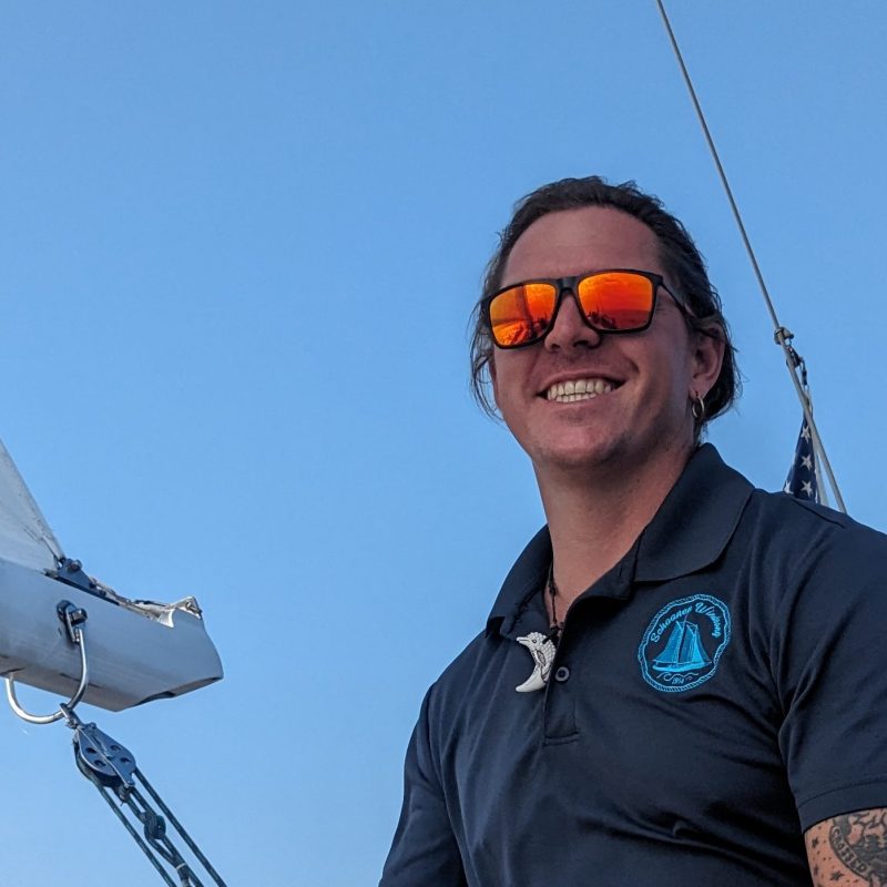 Person with tattoos steering a sailboat, wearing sunglasses and a dark shirt on a sunny day.