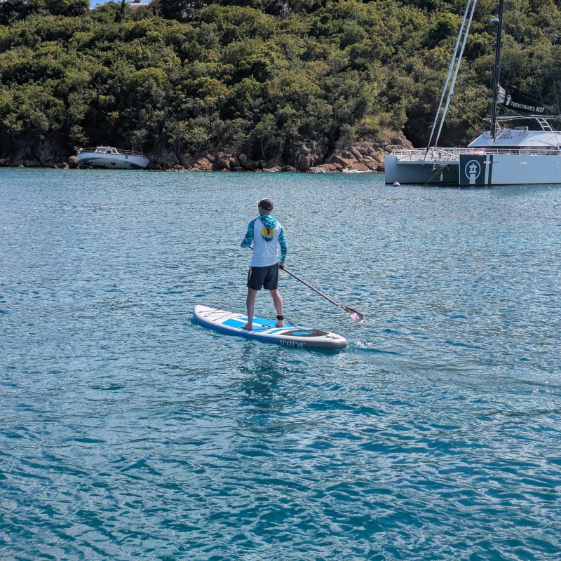 Person paddleboarding on calm water near a yacht and lush shore.